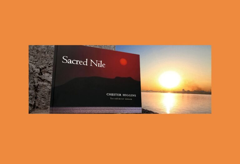 A book titled "Sacred Nile" by Chester Higgins rests upright on a stone surface. Behind it is a calm river with the sun setting near the horizon, casting a warm golden glow over the water and sky. The background and border are solid orange.