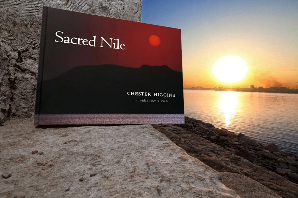 A coffee table book titled "Sacred Nile" by Chester Higgins with text by Betsy Kissam rests on a stone surface next to a calm river. In the background, the sun is setting, casting a warm glow over the water and the distant shoreline. The cover shows a dark landscape with a red sun.