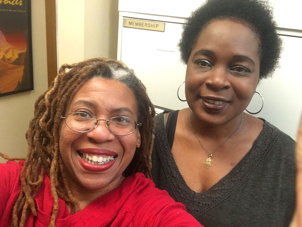 Two women smile at the camera indoors. One, in a red top with glasses and long, highlighted dreadlocks, takes the selfie. The other, wearing a dark V-neck sweater and hoop earrings, stands beside her. They look happy. A “Membership” sign is visible in the background.