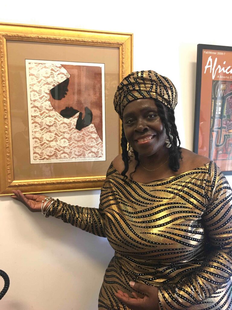 A smiling woman wearing a matching gold and black patterned headwrap and dress stands beside a framed artwork. The artwork features a side profile silhouette on a textured background. She holds the frame with one hand, and the wall behind contains another framed picture.