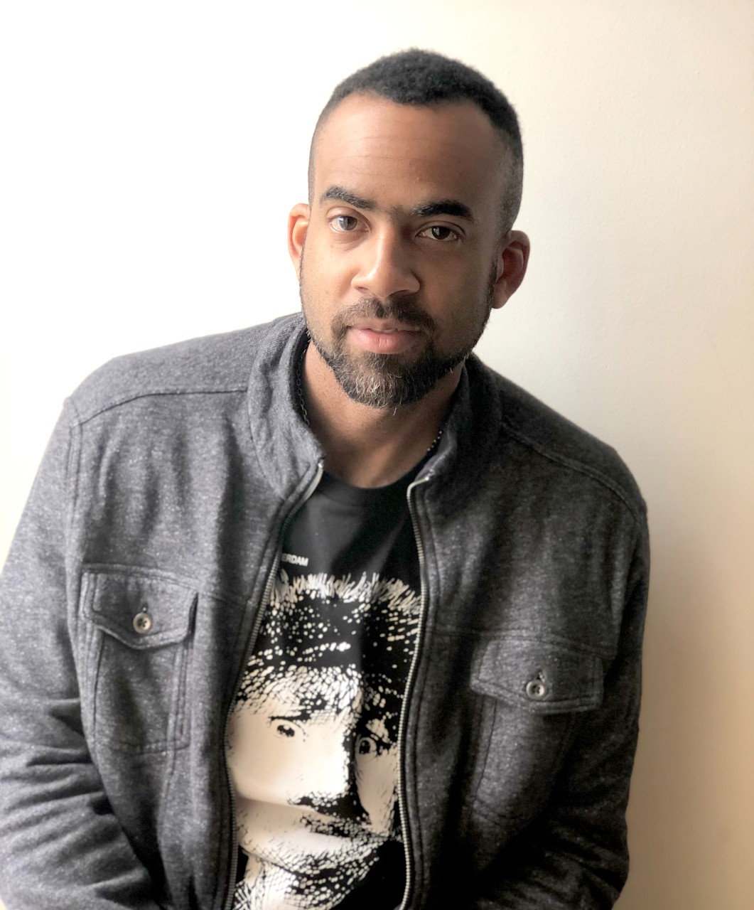 A man with a short beard and mustache is seen against a plain white background. He is wearing a dark gray jacket with buttoned pockets over a black T-shirt featuring a white graphic design of a face. The man has a neutral expression and short, closely-cropped hair.