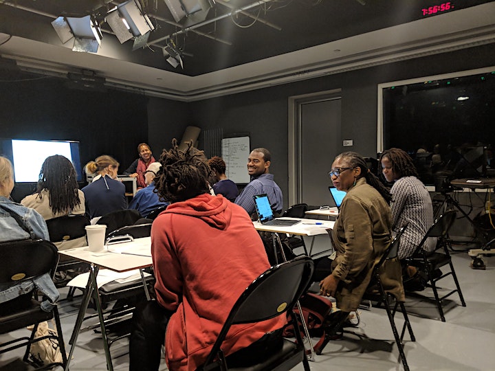 A diverse group of people are seated in a classroom setting, facing a woman standing near a whiteboard. The room is dimly lit, with a large screen and studio lights in the background. Participants are engaged, with some looking at laptops and others taking notes.