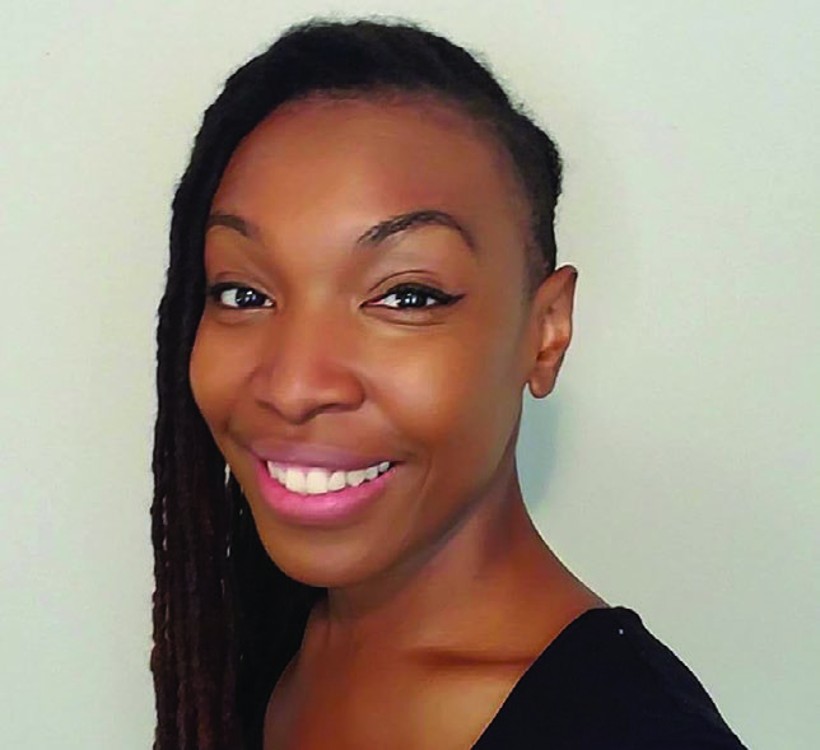 A person with long, dark braided hair and a shaved portion on the side smiles warmly at the camera. They are wearing a black top and have a neutral background behind them. They have smooth skin, well-defined eyebrows, and are slightly tilted to one side for the photo.