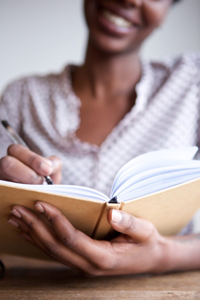 A person with a blurred smiling face writes in an open spiral notebook with a pen. They're wearing a patterned blouse. The focus is on the hands and notebook, creating a sense of engagement with writing. The background is softly blurred, emphasizing the activity.