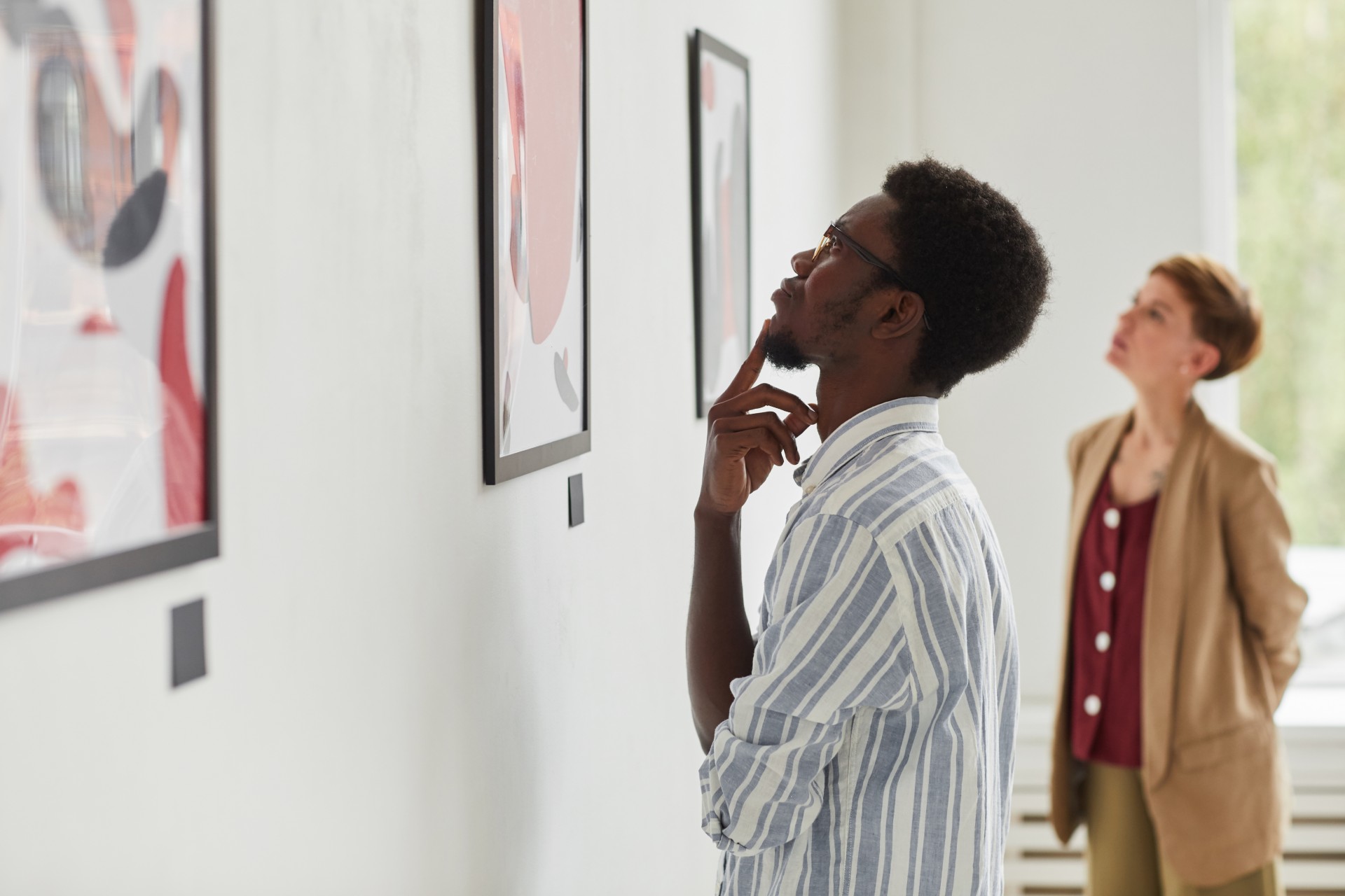 A man in a striped shirt thoughtfully observes artwork on a gallery wall. A woman in a beige blazer stands slightly behind him, also looking at the art. The room is bright, with several framed abstract pieces lining the white wall.