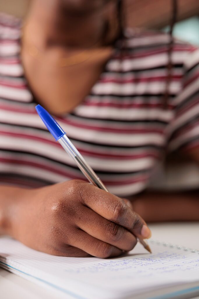 A person with braided hair writes in a notebook with a blue pen. They wear a striped shirt with horizontal lines in black, white, and red. The focus is on the hand holding the pen and the notebook with cursive writing, while the background is softly blurred.