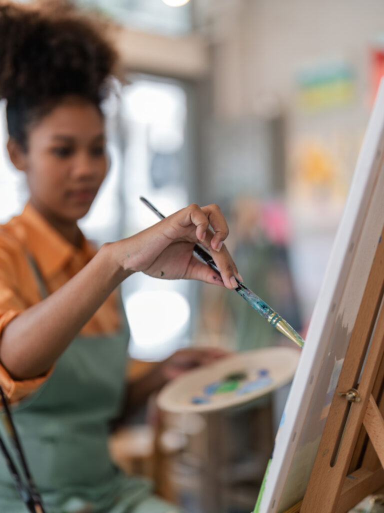 A woman with curly hair, wearing an orange shirt and green apron, is painting on a canvas placed on an easel. She holds a paintbrush in her right hand and a palette with various colors in her left hand. The background is softly blurred, suggesting an art studio setting.