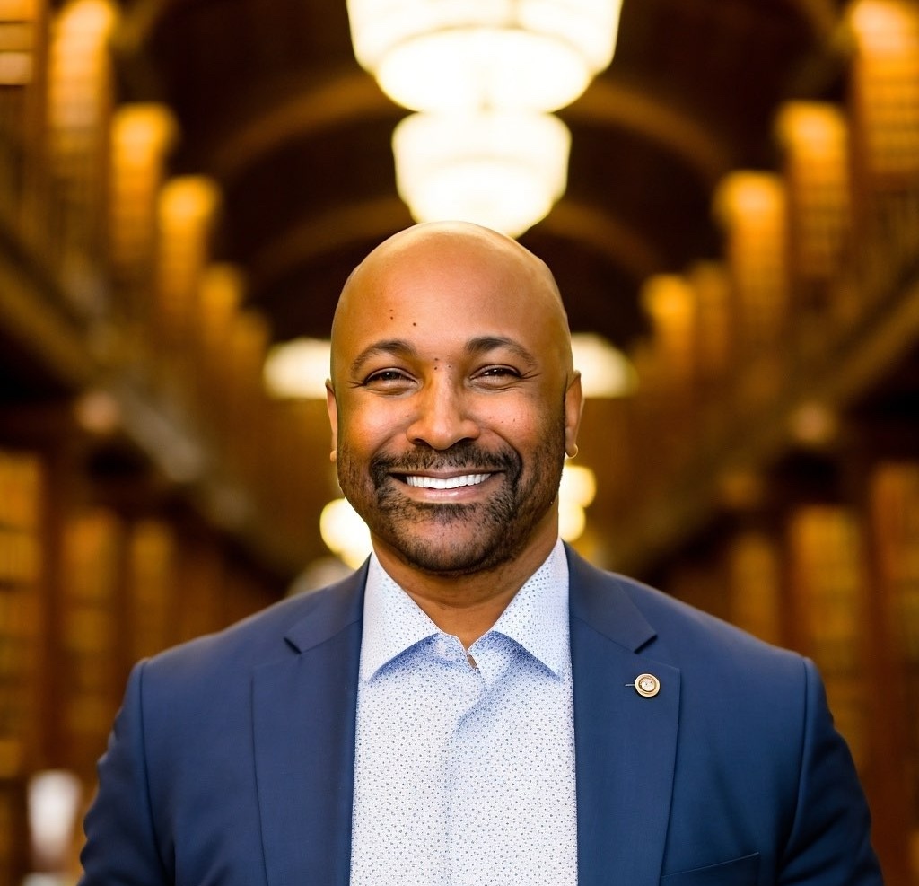 A bald man with a beard, wearing a blue suit jacket and white shirt, smiles at the camera. He stands in a grand library with tall bookshelves and arched ceiling, warmly lit by large chandeliers in the background.
