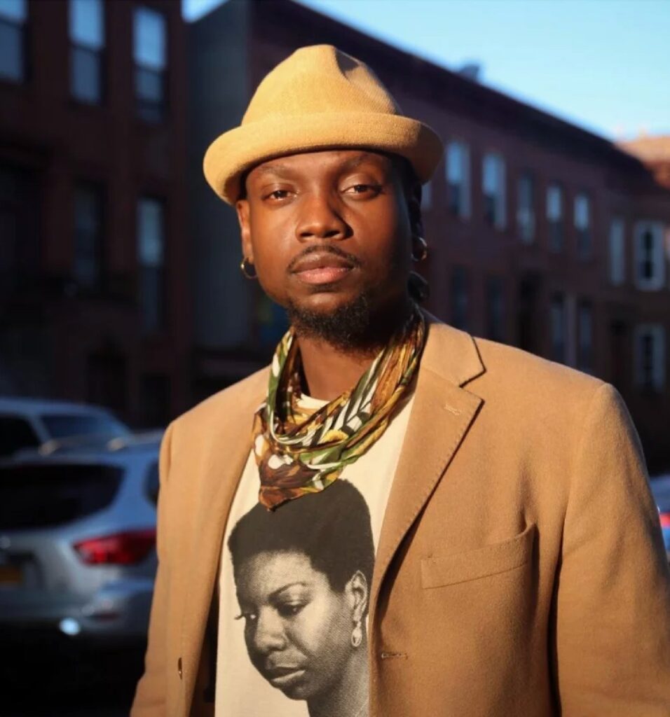 A stylish man stands outdoors in warm sunlight, wearing a tan blazer, a beige hat, a patterned scarf, and a T-shirt featuring a black-and-white portrait of a woman. Parked cars and brownstone buildings are visible in the blurred background.