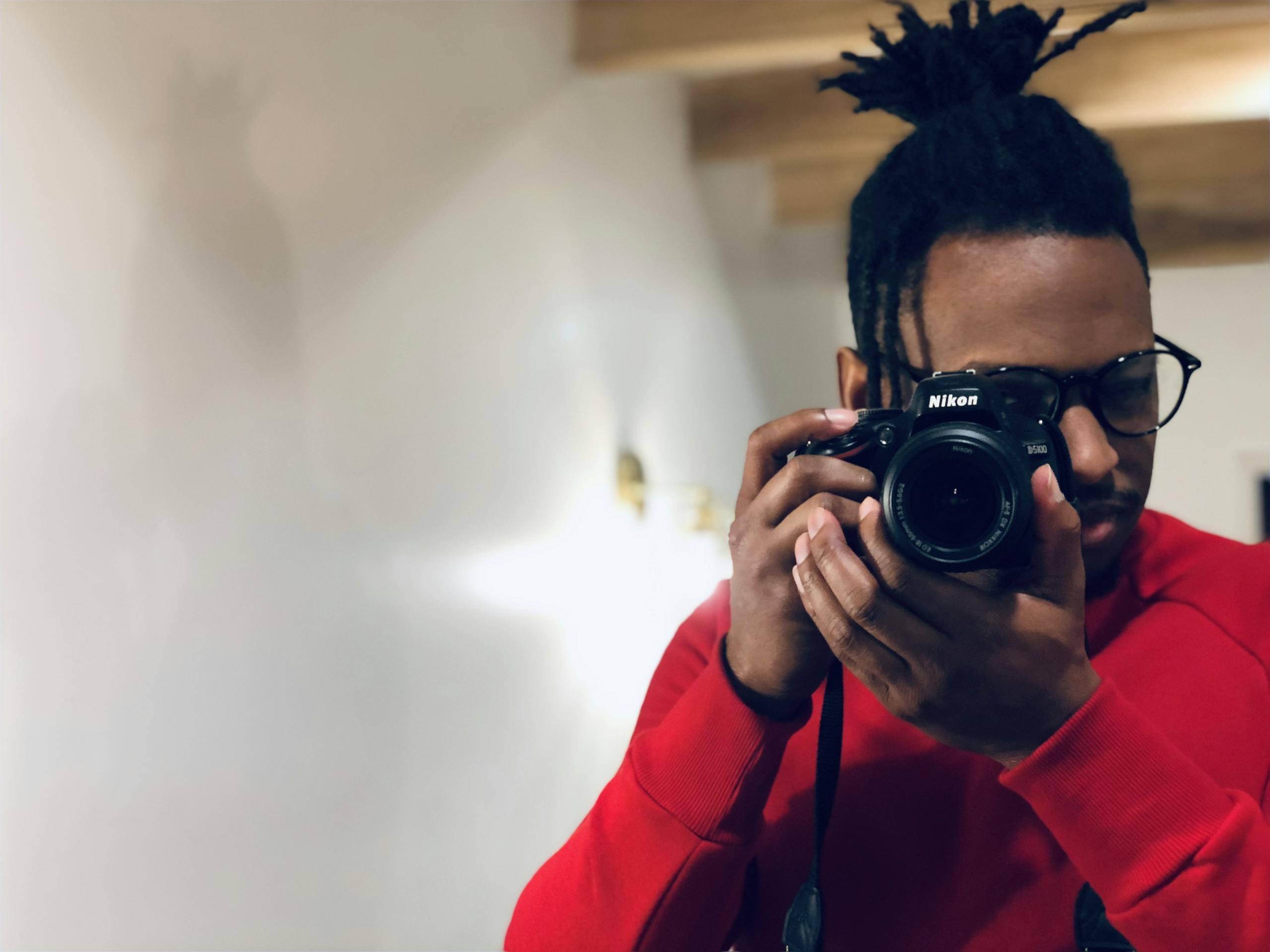 A man in red sweater holds a professional camera indoors, capturing a self-portrait.