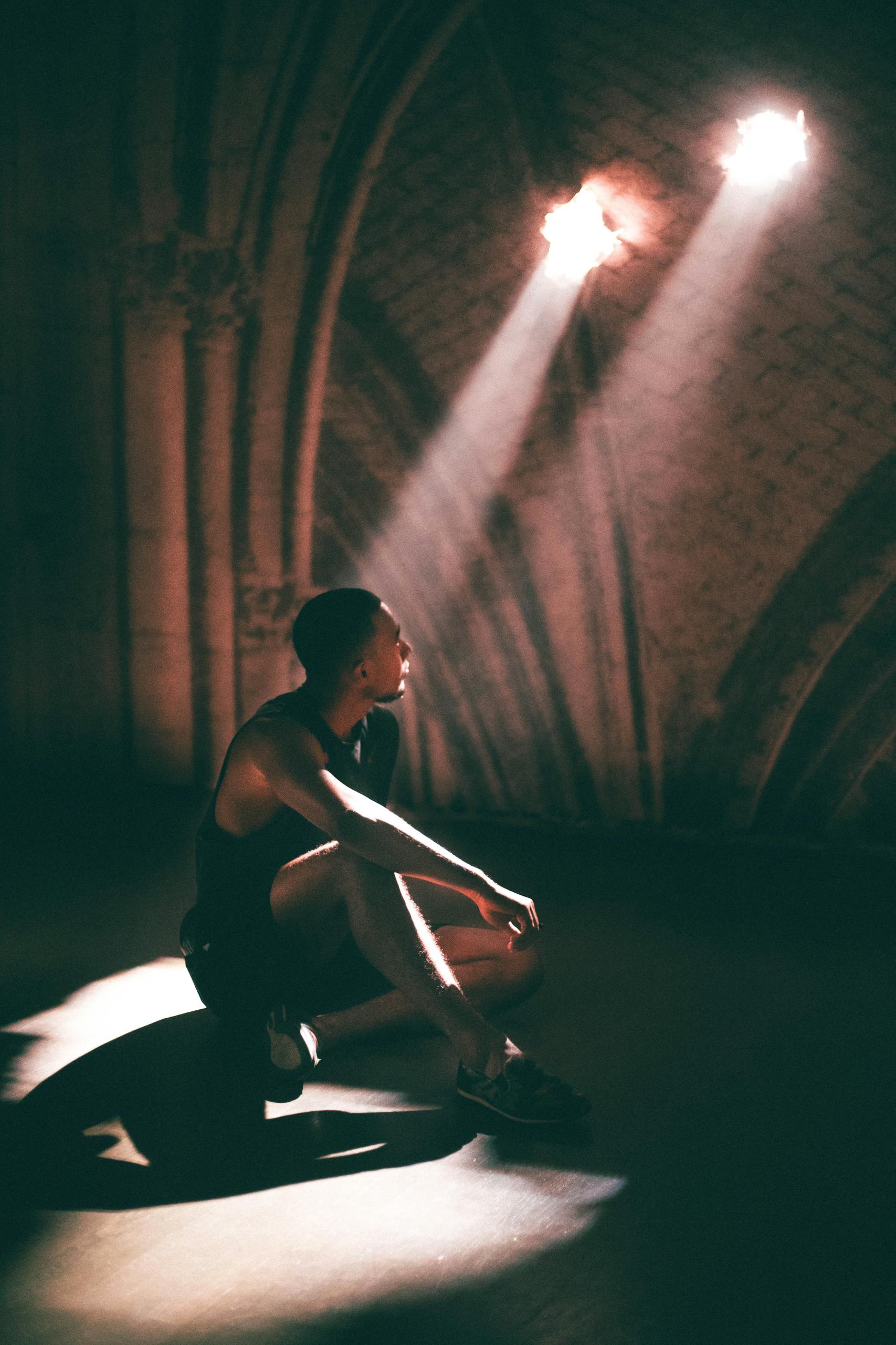 A young man sits in dramatic lighting under spotlight beams in a theater setting.